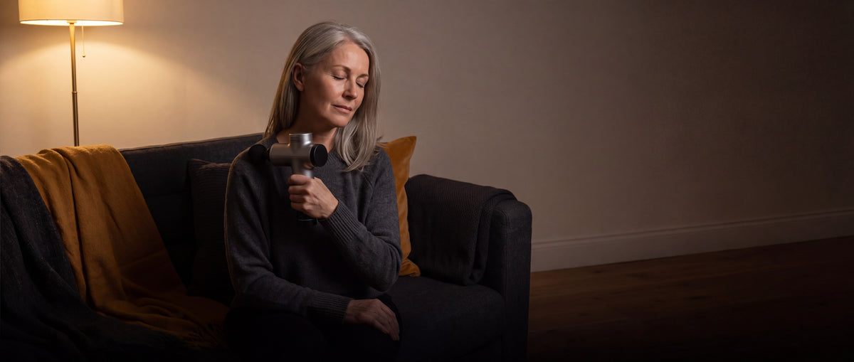 A woman in her fifties easing her shoulder with a massage gun in evening light